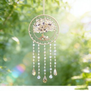 May include: A handcrafted dream catcher featuring a golden wire frame with a tree design. The tree is adorned with white and purple beads, and the dream catcher has dangling strands of clear and amber-colored crystals. The background is a blurred green.