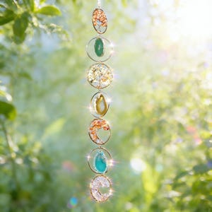 May include: A hanging suncatcher with seven circular and oval-shaped sections. Each section features a different colored stone or crystal, including orange, green, and clear. The sections are connected by gold-colored metal rings and chains. The background is a blurred green.