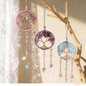 Puede incluir: Tres atrapasueños colgantes con un diseño de árbol de la vida. Cada uno presenta un marco circular con un árbol de alambre y piedras de colores. Los atrapasueños son de color rosa, morado y azul, con cuentas y dijes de cristal colgantes.