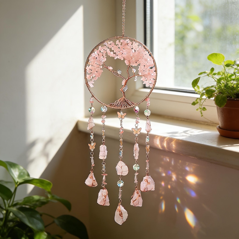 May include: A rose gold metal tree of life dream catcher with pink crystal accents. The circular design features a detailed tree with pink crystal leaves and hanging strands of crystals, butterflies, and raw rose quartz stones.