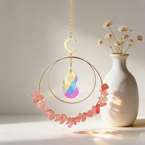 May include: A gold-colored suncatcher with a teardrop-shaped crystal prism and a crescent moon charm. The suncatcher features a circular design adorned with pink gemstones. A white vase with dried flowers is in the background.