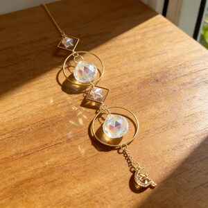 May include: A gold-colored suncatcher with a celestial theme. It features two clear, faceted crystal balls suspended within gold rings, connected by gold square frames with star accents. A crescent moon and star charm dangles at the bottom.