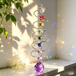 Può includere: Un carillon a vento a spirale in metallo argentato con una fila verticale di cristalli di vetro sfaccettati multicolori trasparenti, rossi, gialli, verdi, blu e viola, che termina con una grande sfera di cristallo viola che riflette la luce.