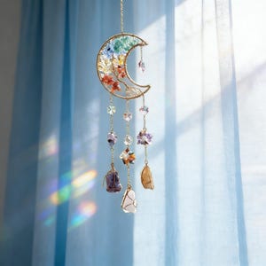 May include: A gold-colored crescent moon-shaped suncatcher with colorful crystals and gemstones. The moon is filled with various colored crystals and has dangling strands of crystals and gemstones. The suncatcher hangs in front of a blue curtain.