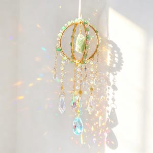 May include: A decorative suncatcher with a gold-colored metal frame, adorned with green stones and clear crystal pendants. The suncatcher features a large central crystal and smaller dangling crystals, creating a prism effect with light.