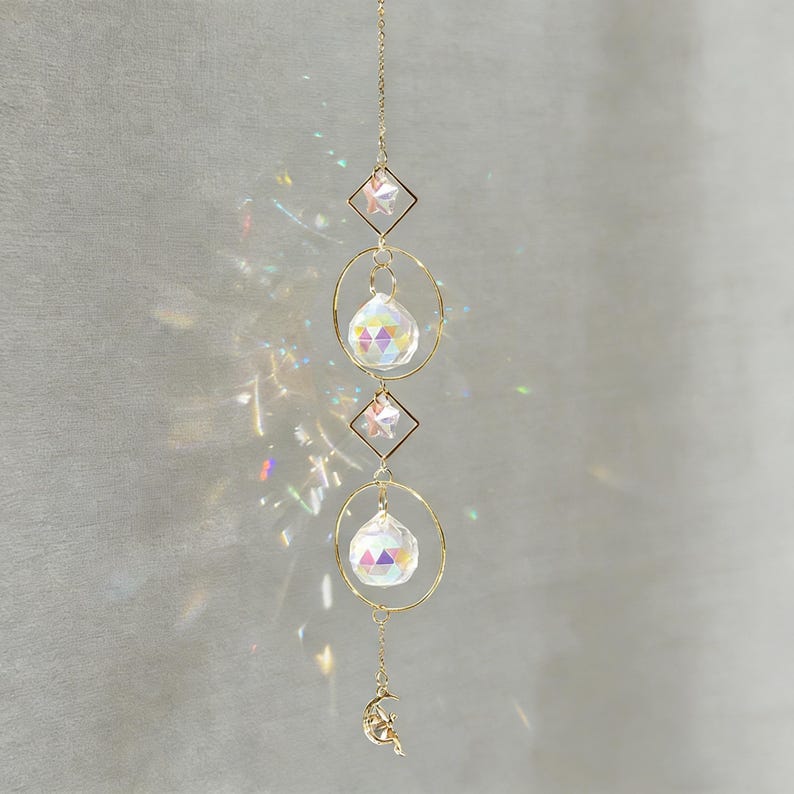 May include: A decorative suncatcher with a gold chain, featuring three clear crystal spheres, two star-shaped accents, and a crescent moon charm. The suncatcher is designed to catch and reflect light.