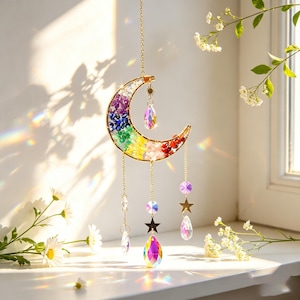 May include: A crescent moon suncatcher featuring a gold-toned wire frame filled with small, multi-colored gemstone chips in a rainbow gradient. Three gold chains hang from the bottom, each adorned with a clear, iridescent crystal teardrop and a small gold star charm.