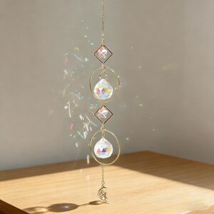 May include: A decorative suncatcher with a gold chain, geometric shapes, and crystal prisms. The suncatcher features two diamond-shaped frames with star accents, two crystal spheres, and a crescent moon charm at the bottom. The prisms cast colorful light reflections.