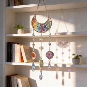 Pu&ograve; includere: Un acchiappasole a tema celeste con una luna crescente dorata piena di pietre colorate. Dalla luna pendono ornamenti a forma di stella e gemma e cristalli a forma di goccia. L'acchiappasole &egrave; progettato per essere appeso e catturare la luce.