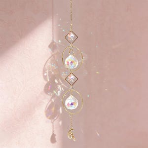 Op de afbeelding: Een hangende kristallen zonnevanger met een gouden ketting, diamant- en cirkelaccenten en een maansikkelsieraad. De zonnevanger heeft twee kristallen bollen en twee stervormige kristallen. De achtergrond is zachtroze.
