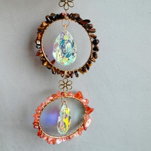 May include: A two-tiered suncatcher with a gold-coloured wire frame. The top circle is adorned with brown stones and a teardrop-shaped crystal. The bottom circle features orange stones and a similar crystal. Floral accents connect the tiers.