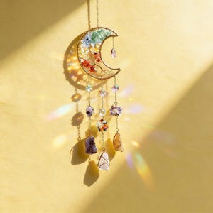 May include: A crescent moon-shaped suncatcher with a gold-colored wire frame. The moon is filled with colorful crystals and adorned with hanging gemstones and crystal accents. The suncatcher is suspended by a delicate chain.