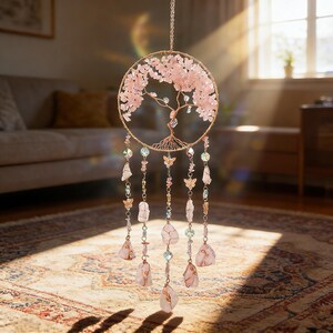 Puede incluir: Un atrapasue&ntilde;os de &aacute;rbol de la vida de cuarzo rosa. El marco circular es de alambre dorado, con piedras rosas que forman las hojas del &aacute;rbol. Cuelgan debajo hilos de cristales, cuentas y dijes de mariposas. El atrapasue&ntilde;os cuelga frente a una ventana.