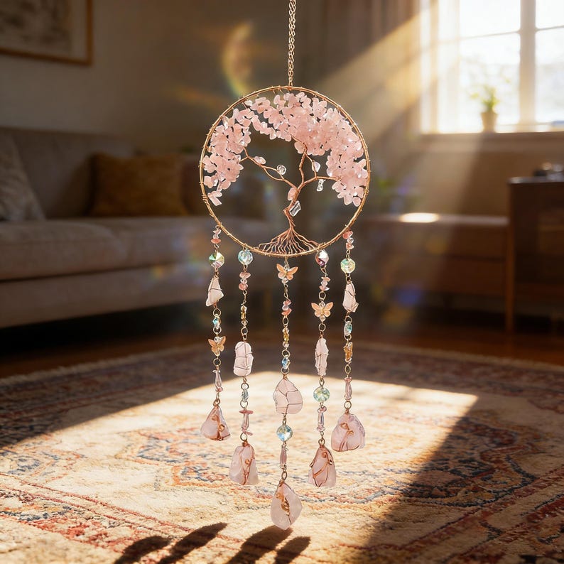 Puede incluir: Un atrapasue&ntilde;os de &aacute;rbol de la vida de cuarzo rosa. El marco circular es de alambre dorado, con piedras rosas que forman las hojas del &aacute;rbol. Cuelgan debajo hilos de cristales, cuentas y dijes de mariposas. El atrapasue&ntilde;os cuelga frente a una ventana.