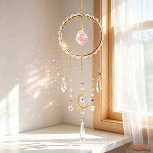 May include: A gold-toned metal hoop suncatcher featuring a central raw pink crystal, clear glass beads, and hanging chains. The design includes a gold crescent moon charm, small purple accents, and multiple clear glass prisms that refract light.