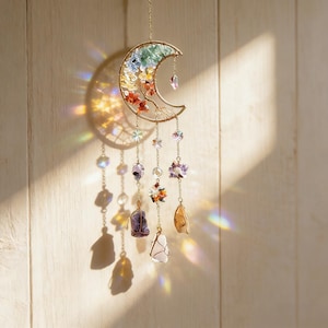 May include: A gold-colored crescent moon suncatcher with colorful crystals and gemstones. The moon is adorned with a rainbow of crystals and small floral accents. Dangling below are various gemstones and crystal beads, creating a decorative and spiritual piece.
