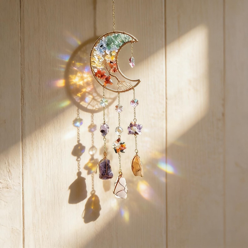 May include: A gold-colored crescent moon suncatcher with colorful crystals and gemstones. The moon is adorned with a rainbow of crystals and small floral accents. Dangling below are various gemstones and crystal beads, creating a decorative and spiritual piece.