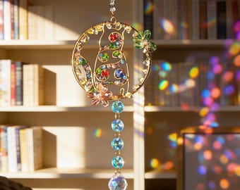 May include: A decorative suncatcher with a gold-coloured wire cat design, embellished with colourful gemstones and floral details. A strand of blue and clear crystal beads hangs below, ending in a faceted crystal ball. The suncatcher is suspended by a delicate gold chain.