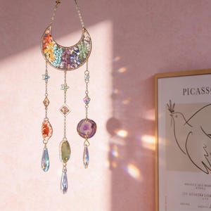 Pu&ograve; includere: Un acchiappasole a tema celeste con una luna a mezzaluna piena di pietre colorate. Catene dorate sospendono cristalli a forma di stella e gemma, creando un pezzo decorativo e accattivante. L'acchiappasole &egrave; progettato per catturare e riflettere la luce.
