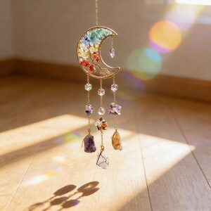 May include: A gold-colored crescent moon-shaped wall hanging with colorful gemstones and crystals. The moon features a rainbow of colors and is adorned with hanging crystals and floral accents. The decorative piece is suspended by a delicate chain.