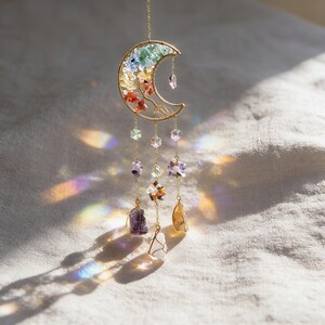 May include: A gold-colored crescent moon suncatcher with colorful gemstone accents. The moon features a rainbow of small stones and dangling crystals. The suncatcher hangs from a delicate chain and has several strands of crystals and gemstones.
