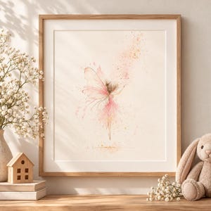 May include: A framed wall art print featuring a watercolour illustration of a fairy with delicate pink wings and a matching dress, surrounded by soft pink and gold paint splatters on a cream background, encased in a light wood frame.