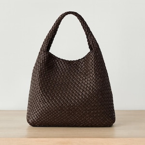 May include: A dark brown woven leather shoulder bag with a single handle. The bag has a rounded shape and a textured surface. The bag is sitting on a light brown wooden surface.