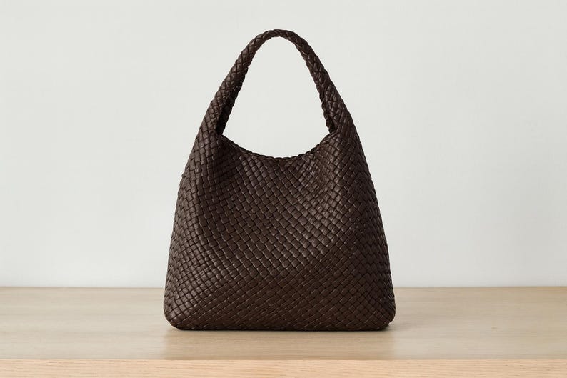 May include: A dark brown woven leather shoulder bag with a single handle. The bag has a rounded shape and a textured surface. The bag is sitting on a light brown wooden surface.