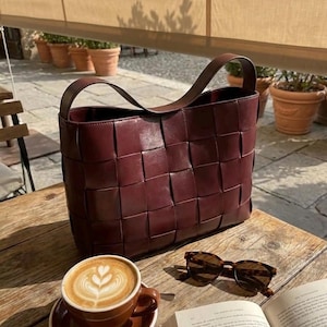 Op de afbeelding: Een bordeauxrode leren tote bag met een geweven design staat op een houten tafel. Daarnaast staan een kop koffie met latte art, een zonnebril en een open boek. De tas heeft een enkele handgreep.