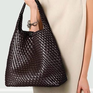 May include: A dark brown woven leather tote bag with a wide shoulder strap. The bag has a large capacity and a rectangular shape. The woven pattern creates a textured surface. The bag is held by a person wearing a cream-colored sleeveless dress.