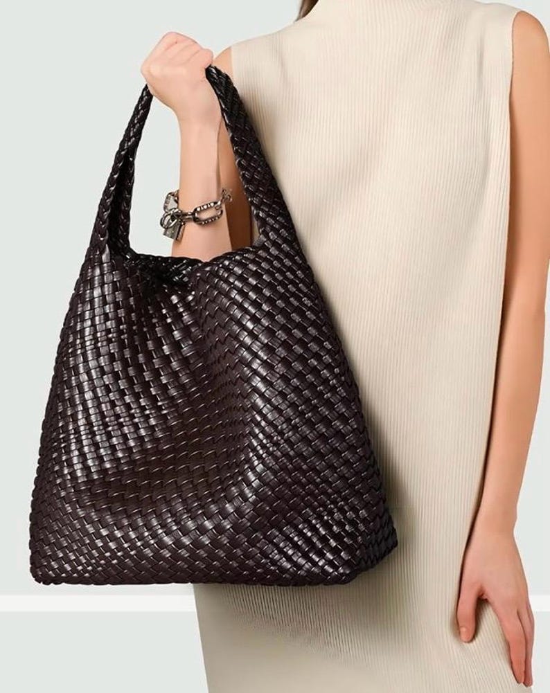 May include: A dark brown woven leather tote bag with a wide shoulder strap. The bag has a large capacity and a rectangular shape. The woven pattern creates a textured surface. The bag is held by a person wearing a cream-colored sleeveless dress.