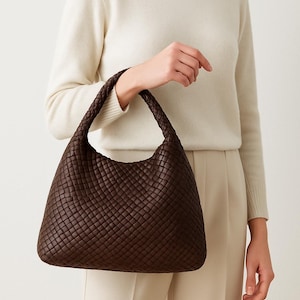 May include: A dark brown woven leather shoulder bag with a braided handle. The bag has a rounded shape and a textured surface. The bag is held by a person wearing a cream-colored sweater and pants.