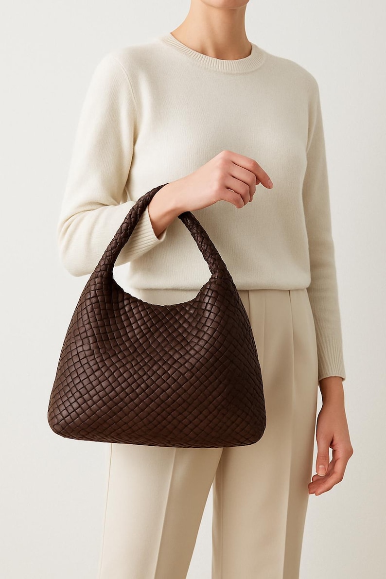 May include: A dark brown woven leather shoulder bag with a braided handle. The bag has a rounded shape and a textured surface. The bag is held by a person wearing a cream-colored sweater and pants.