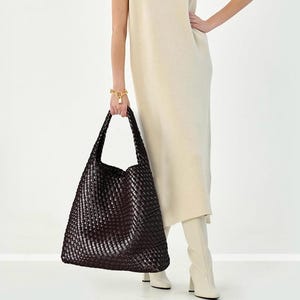 May include: A large, dark brown woven tote bag with long handles. The bag is held by a person wearing a cream-colored dress and white boots. A gold bracelet with a lock charm is visible on the person's wrist.