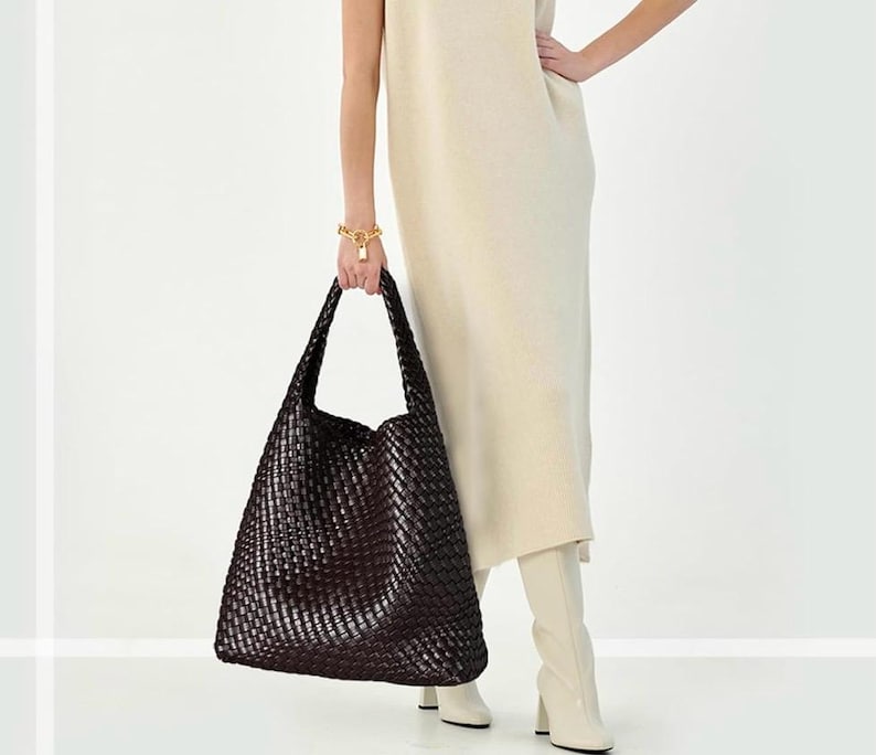 May include: A large, dark brown woven tote bag with long handles. The bag is held by a person wearing a cream-colored dress and white boots. A gold bracelet with a lock charm is visible on the person's wrist.