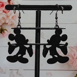 K&ouml;nnte beinhalten: Schwarze Silhouette-Ohrringe von Mickey Mouse, die an einem schwarzen Ausstellungsst&auml;nder h&auml;ngen. Die Ohrringe zeigen die ikonische Form der Maus, einschlie&szlig;lich ihrer gro&szlig;en Ohren, ihres runden Gesichts und ihrer behandschuhten H&auml;nde. Der Hintergrund zeigt ein verschwommenes Blumenarrangement.