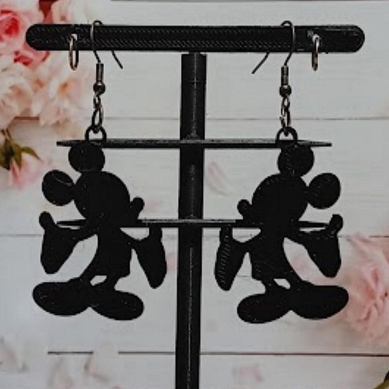 K&ouml;nnte beinhalten: Schwarze Silhouette-Ohrringe von Mickey Mouse, die an einem schwarzen Ausstellungsst&auml;nder h&auml;ngen. Die Ohrringe zeigen die ikonische Form der Maus, einschlie&szlig;lich ihrer gro&szlig;en Ohren, ihres runden Gesichts und ihrer behandschuhten H&auml;nde. Der Hintergrund zeigt ein verschwommenes Blumenarrangement.