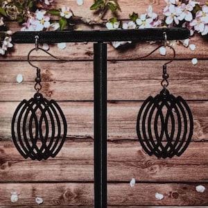 May include: A pair of black, geometric earrings with a layered, oval design. The earrings are suspended from a black metal hook and are displayed on a black stand against a wooden background with floral accents.