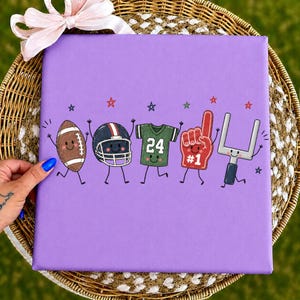 May include: A square purple canvas featuring a cartoon illustration of five American football-themed characters with smiling faces, including a brown football, a black helmet, a green jersey with the number 24, a red foam finger, and a goalpost.