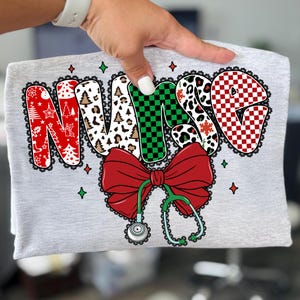 Peut inclure: Un t-shirt gris avec le mot "NURSE" dans un design festif. Les lettres sont d&eacute;cor&eacute;es de motifs de No&euml;l, d'un n&oelig;ud rouge et d'un st&eacute;thoscope. Le design comprend des couleurs rouge, vert, blanc et noir, avec une bordure en dentelle.