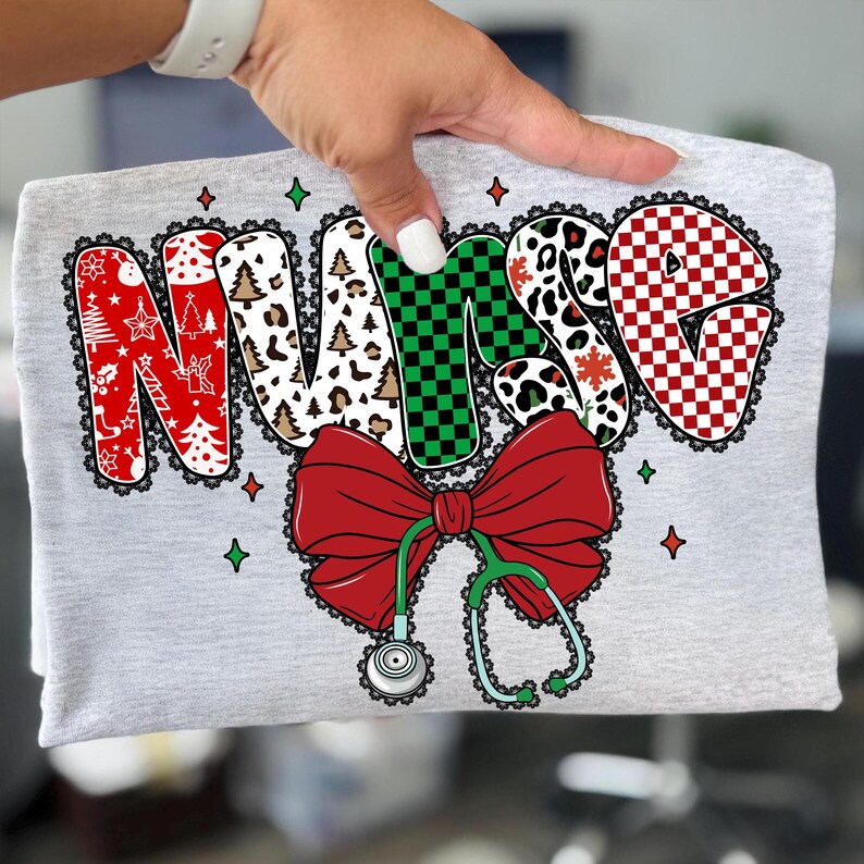 Peut inclure: Un t-shirt gris avec le mot "NURSE" dans un design festif. Les lettres sont d&eacute;cor&eacute;es de motifs de No&euml;l, d'un n&oelig;ud rouge et d'un st&eacute;thoscope. Le design comprend des couleurs rouge, vert, blanc et noir, avec une bordure en dentelle.