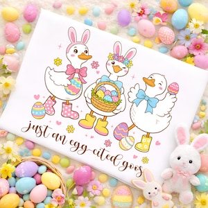 May include: White t-shirt featuring three cartoon geese dressed for Easter. The geese wear bunny ears, bows, and boots, and carry Easter eggs and a basket. The text "just an egg-cited goose" is written in script.