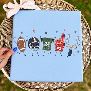 May include: A light blue square fabric item featuring a cartoon graphic of a brown football, a dark helmet, a green jersey with the number 24, a red foam finger with the number 1, and a goalpost, all with arms, legs, and small stars.