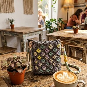 May include: A gray crocheted crossbody bag with a floral pattern in pastel colors, including yellow, pink, and green. The bag has a braided strap with yellow beads and a tassel. The bag is sitting on a wooden table next to a potted plant and a cup of coffee.