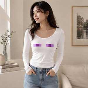 Op de afbeelding: Een witte longsleeve met een ronde hals en een ontwerp van twee paarse rechthoeken. De top wordt gecombineerd met een blauwe jeans. Het model draagt de top.