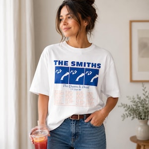 May include: White t-shirt featuring the band name "THE SMITHS" in blue block letters above a blue rectangle with a graphic and the text "The Queen Is Dead US Tour 86". The shirt is worn with blue jeans.