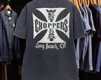 May include: Dark gray t-shirt with a large white graphic on the back. The graphic features a cross design with the words "WEST COAST CHOPPERS" and "Long Beach, CA" in a bold, stylized font. The shirt is hanging on a wooden hanger.