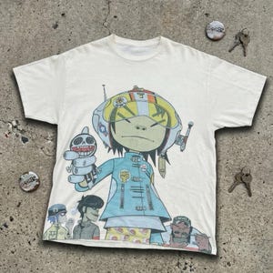 Könnte beinhalten: Weißes T-Shirt mit einem farbenfrohen Cartoon-Motiv eines Charakters in blauer Uniform und Helm, der ein Spielzeug hält. Die Grafik enthält weitere Charaktere und das Wort "OWE". Das T-Shirt liegt auf einer Betonoberfläche.