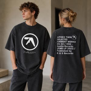 May include: Two people wearing dark gray t-shirts. The front of the shirt has a white circular logo with a stylized design and the text "Aphex Twin". The back of the shirt has the text "Aphex Twin Selected Ambient Works 85-92 / 1992 - Apollo Records (AMB LP 3922) / Published by R & S Records."