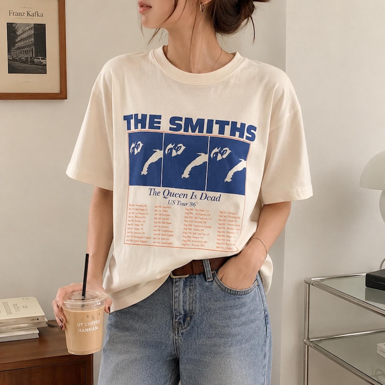 May include: Cream-colored t-shirt featuring the band name "THE SMITHS" in blue block letters above a blue rectangle with a graphic and the text "The Queen Is Dead US Tour '86". The shirt has short sleeves and a crew neck.
